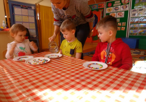 Dzieci przyglądają się jak pani Ewelina nalewa wodę na talerzyk jednego z chłopców.